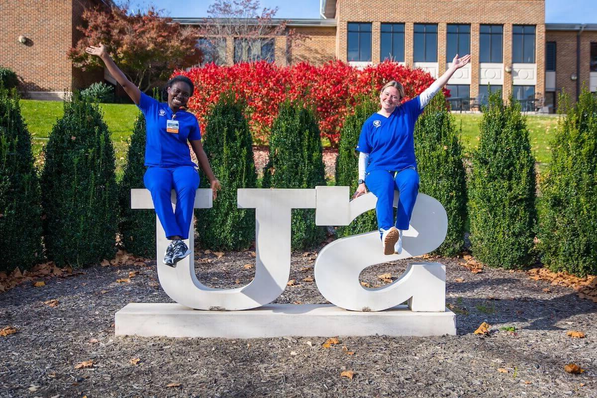 Two Shenandoah nursing students, in blue scrubs, 秋天坐在澳门网上赌博平台广场上的SU雕像上.